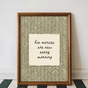 Op de afbeelding: Een ingelijste wanddecoratie met een donker olijfgroen en crème visgraatpatroon. In het midden bevindt zich een crèmekleurig papier met rafelige randen, waarop de zwarte cursieve tekst staat: his mercies are new every morning.