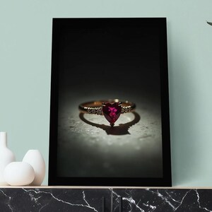 Puede incluir: Un anillo de oro con una piedra preciosa roja oscura en forma de corazón. El anillo se muestra en un marco negro, proyectando una sombra. El anillo está sobre un mueble con vetas de mármol, con jarrones decorativos blancos. El fondo es una pared verde claro.