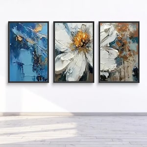 Puede incluir: Tres obras de arte abstractas enmarcadas. El marco izquierdo presenta pinceladas azules y doradas. El marco central muestra una flor blanca con un centro amarillo. El marco derecho tiene pinceladas blancas, marrones y azules. Todos los marcos tienen bordes negros.