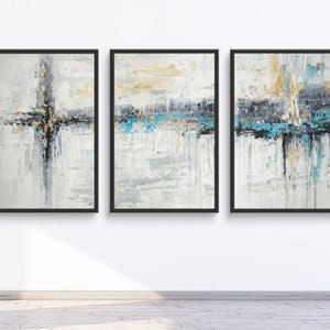 Puede incluir: Tres pinturas abstractas enmarcadas con marcos negros. La obra de arte presenta una mezcla de colores, incluyendo blanco, gris, negro, dorado y turquesa. Las pinturas se exhiben en una pared blanca sobre un suelo de madera.