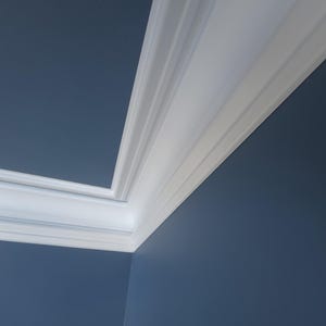 Puede incluir: Una esquina de habitación con moldura blanca contra una pared azul oscuro. La moldura tiene un diseño detallado y en capas, contrastando con la superficie lisa de la pared. La imagen resalta detalles arquitectónicos.