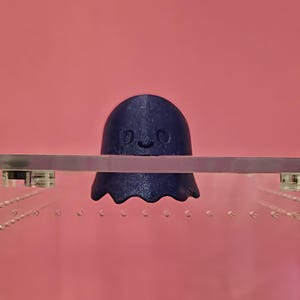 May include: A navy blue ghost-shaped decorative object with a smiling face. The ghost is positioned above a clear, rectangular container with a pink background. The object appears to be 3D-printed.