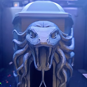 May include: A 3D-printed sculpture of a snake's head with an open mouth, fangs, and a forked tongue. The sculpture is primarily blue-gray and has a detailed, textured surface. The snake's head is the focal point of the artwork.