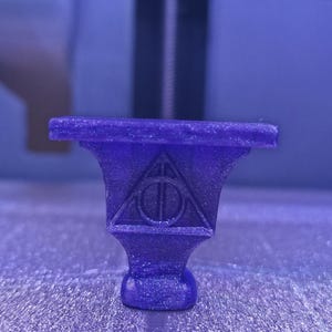 May include: A 3D-printed, purple, decorative object featuring the Deathly Hallows symbol. The object has a pedestal base and a flat top. The Deathly Hallows symbol is a triangle, a circle, and a vertical line.