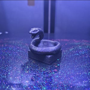 May include: A dark grey snake figurine coiled around a small, circular base. The snake has an open mouth. It is set on a dark, reflective surface with colourful specks, under a blue light.