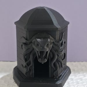 Könnte beinhalten: Eine schwarze, 3D-gedruckte dekorative Box mit gewölbtem Deckel und einem Medusa-ähnlichen Gesicht, das in die Vorderseite geschnitzt ist. Die Box hat eine rechteckige Basis und eine kleine Öffnung. Das Gesamtdesign ist detailliert und strukturiert.
