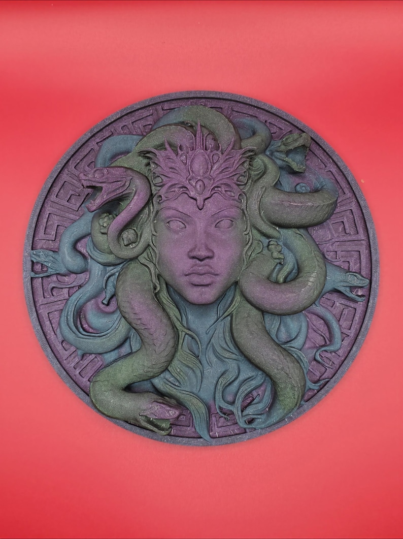 May include: A circular relief sculpture of Medusa with snakes for hair, in shades of purple, green, and blue. The Medusa figure has a crown and is set against a patterned border. The background is a solid red.