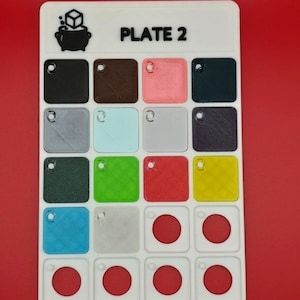 May include: A white plastic game board labeled "PLATE 2" with a variety of colored squares and circles. The board features a logo of a cauldron with a cube above it. The squares are in various colors including black, brown, pink, and green.