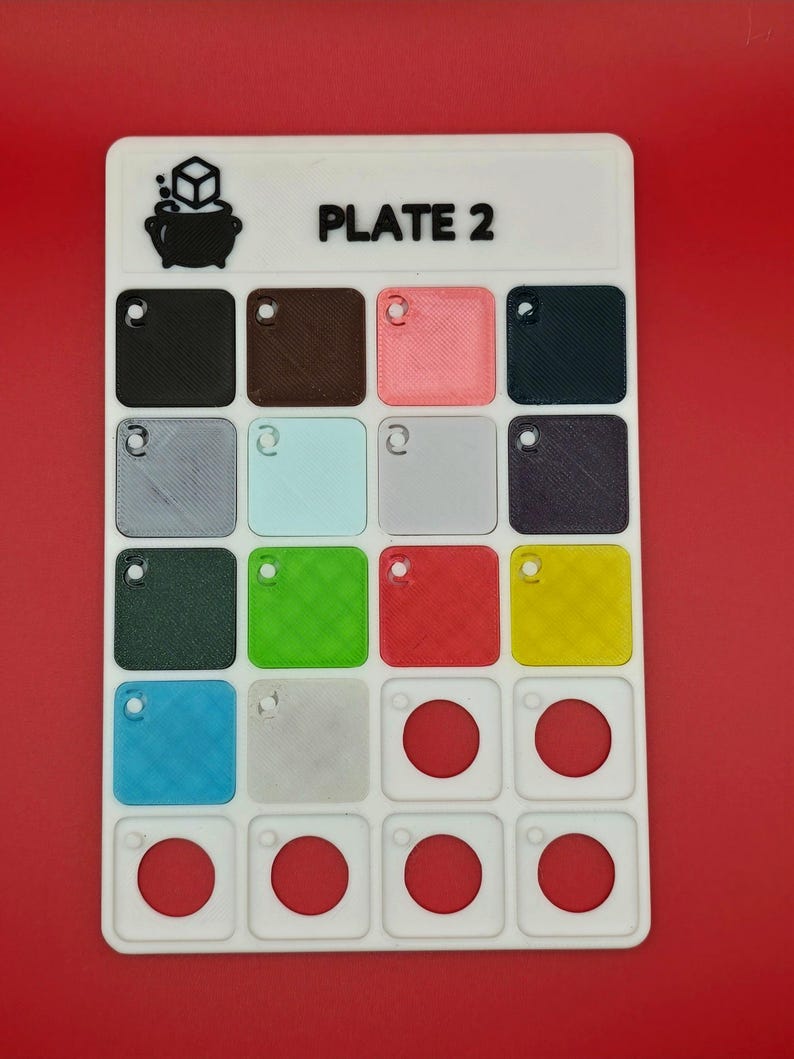 May include: A white plastic game board labeled "PLATE 2" with a variety of colored squares and circles. The board features a logo of a cauldron with a cube above it. The squares are in various colors including black, brown, pink, and green.
