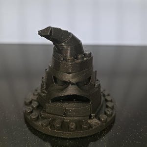 May include: A black 3D-printed Sorting Hat, a fictional character from the Harry Potter series. The hat has a wide brim, a conical shape, and a slightly bent top. The hat's surface has a textured appearance.