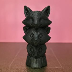 Könnte beinhalten: Eine dunkelgraue Totempfahlskulptur mit einem Fuchs, einem Waschbären und einer Eule, die übereinander gestapelt sind. Die Skulptur ist 3D-gedruckt und steht vor einem rosa Hintergrund. Der Fuchs hat spitze Ohren und ein lächelndes Gesicht.