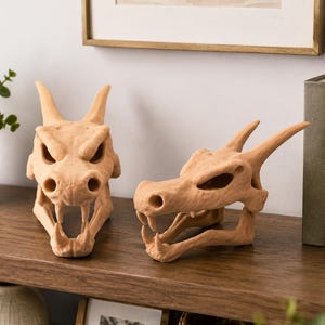 Puede incluir: Dos esculturas de calaveras de dragón beige con detalles, incluyendo cuernos, cuencas de los ojos y mandíbulas abiertas. Las calaveras se exhiben en una estantería de madera, sugiriendo un tema decorativo o artístico.