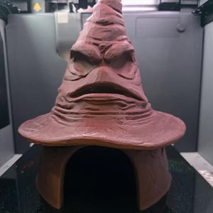 Può includere: Un cappello parlante marrone scolpito, con tesa larga e punta. Il cappello ha una superficie strutturata e dettagliata e un'espressione severa. Il cappello è su una base marrone.