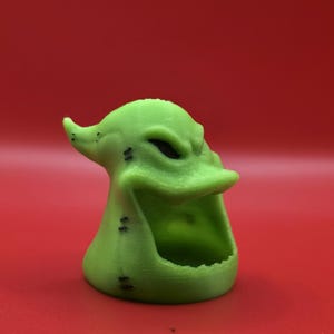 May include: A green, 3D-printed figurine of a character with a wide, toothy grin and black eyes. The figurine has a textured surface and is set against a red background. The character's design includes small black dots.