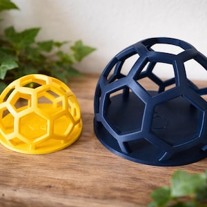 May include: Two decorative, geometric sphere-shaped objects. One is yellow, the other is navy blue. Both have an open, honeycomb-like design. They sit on a wooden surface with green foliage in the background.