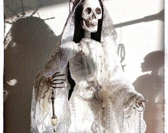 May include: A skeletal figure in a white lace gown and veil, adorned with a halo and holding a scale and a glass jar. The figure has long black hair and a detailed skull. The image is lit with dramatic shadows.