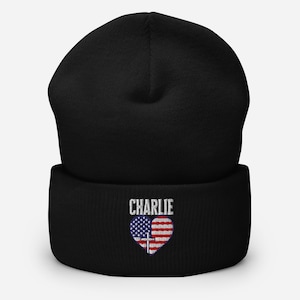 May include: Black beanie with the name "CHARLIE" embroidered in white above a heart-shaped design. The heart features the American flag on the left and red and white stripes with a cross on the right.