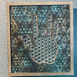 May include: A framed wall art piece with a hand-shaped cutout in the center. The hand is made of a patterned material with a blue and gold color scheme. The background is a similar pattern but in a darker shade of blue.
