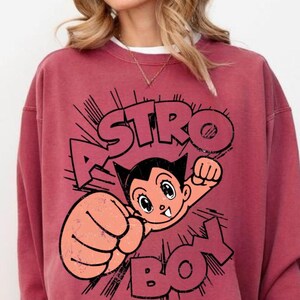 May include: A maroon-colored sweatshirt featuring a graphic of Astro Boy. The design includes the character's image and the words "ASTRO BOY" in a bold, comic-style font. The sweatshirt has a crew neck and long sleeves.
