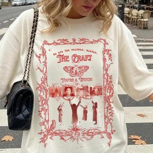 May include: Cream-colored t-shirt with a red graphic from "The Craft" movie. The design includes the movie title, a butterfly, and images of the characters. A beige baseball cap with a black logo is worn. A black purse with a chain strap is also visible.
