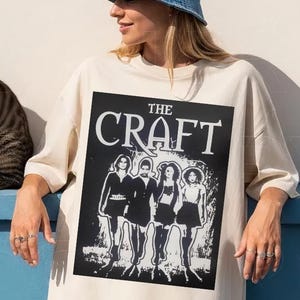 May include: Ivory-colored oversized t-shirt with a black and white graphic from "The Craft" movie. The shirt features the movie title and a stylized image of four women. A denim bucket hat and patterned shorts are also visible.