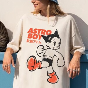 May include: Ivory-colored oversized t-shirt featuring a graphic of Astro Boy with red and black accents. The text "ASTRO BOY" is printed above the character in red, with Japanese text below. A blue denim bucket hat is worn.