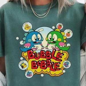 May include: Light green t-shirt featuring a colorful graphic from the video game Bubble Bobble. The design includes two cartoon dinosaurs, bubbles, and the game's logo in yellow and red. The shirt is a short-sleeved crew neck.