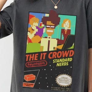 May include: Dark gray t-shirt featuring a graphic from "The IT Crowd." The design includes pixelated characters, the show's title, and the words "Standard Nerds." The shirt has a relaxed fit and short sleeves.