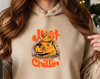 Puede incluir: Sudadera con capucha color arena con bolsillo de canguro y capucha con cordón. La parte delantera presenta un gráfico de un carpincho con un patito de goma y el texto "Just Chillin'" en naranja. La sudadera está hecha de un material suave.
