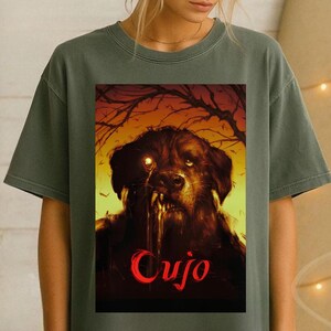 May include: Olive green t-shirt featuring a graphic of a snarling dog with glowing eyes and the word "Cujo" in red. The dog graphic is set against a fiery orange and black background, evoking a horror theme.