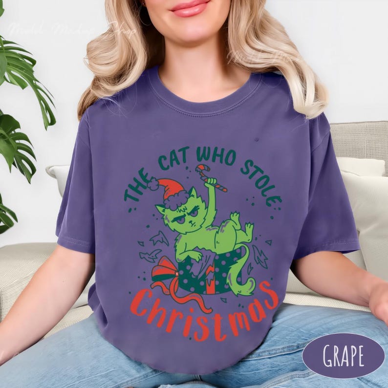 Peut inclure: T-shirt de couleur raisin avec un chat vert portant un bonnet de P&egrave;re No&euml;l et tenant un sucre d'orge. Le texte dit "The Cat Who Stole Christmas". Le chat est dans une bo&icirc;te cadeau avec un n&oelig;ud rouge.