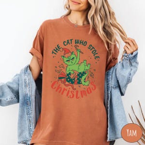 Peut inclure: T-shirt couleur rouille avec un chat vert portant un bonnet de P&egrave;re No&euml;l et tenant un sucre d'orge. Le texte "THE CAT WHO STOLE CHRISTMAS" est imprim&eacute; autour de l'illustration du chat. Le t-shirt a des manches retrouss&eacute;es.