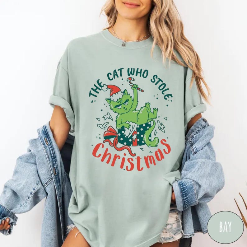 Peut inclure: T-shirt vert clair avec un chat de dessin anim&eacute; portant un bonnet de P&egrave;re No&euml;l et tenant un sucre d'orge. Le texte sur le t-shirt se lit "The Cat Who Stole Christmas" en rouge et vert. Le chat est dans une bo&icirc;te cadeau.