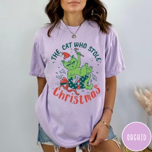 Peut inclure: T-shirt lilas avec un chat vert portant un bonnet de P&egrave;re No&euml;l et tenant un sucre d'orge. Le texte "THE CAT WHO STOLE CHRISTMAS" est imprim&eacute; en vert et rouge.