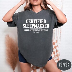 Op de afbeelding: Een antracietgrijs t-shirt met korte mouwen en witte tekst met de tekst CERTIFIED SLEEPMAXXER, gevolgd door SLEEP OPTIMIZATION DIVISION en Est. 2026. Het shirt heeft een ronde hals, een relaxte pasvorm en een vintage gewassen stofafwerking.