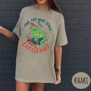Peut inclure: T-shirt kaki avec un chat vert portant un bonnet de P&egrave;re No&euml;l et tenant un sucre d'orge. Le motif indique "The Cat Who Stole Christmas" en vert et rouge. Le chat est assis sur un cadeau emball&eacute;.