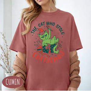 Peut inclure: T-shirt rose poudr&eacute; avec un chat vert portant un bonnet de P&egrave;re No&euml;l et tenant un sucre d'orge. Le texte "THE CAT WHO STOLE CHRISTMAS" est imprim&eacute; en rouge et vert.