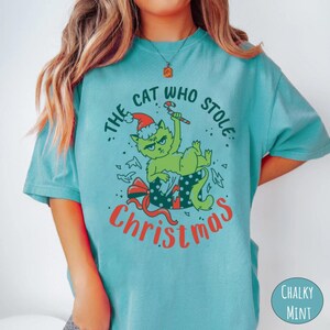 Peut inclure: T-shirt bleu clair avec un motif d'un chat vert portant un bonnet de P&egrave;re No&euml;l et tenant un sucre d'orge. Le texte dit "THE CAT WHO STOLE CHRISTMAS" en rouge et vert. Le chat est dans une bo&icirc;te cadeau.