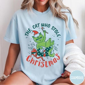 Peut inclure: T-shirt bleu clair avec un chat vert portant un bonnet de P&egrave;re No&euml;l, tenant un sucre d'orge et sortant d'un coffret cadeau. Le texte dit "The Cat Who Stole Christmas" en vert et rouge. Un design de vacances festif.