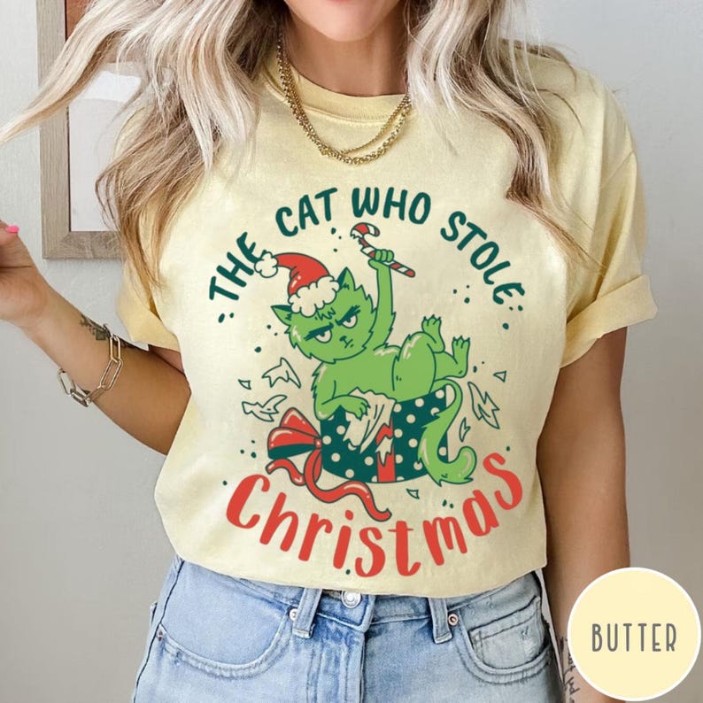 Peut inclure: T-shirt jaune clair avec un motif de chat vert portant un bonnet de P&egrave;re No&euml;l et tenant un sucre d'orge. Le texte "The Cat Who Stole Christmas" est &eacute;crit en vert et rouge.