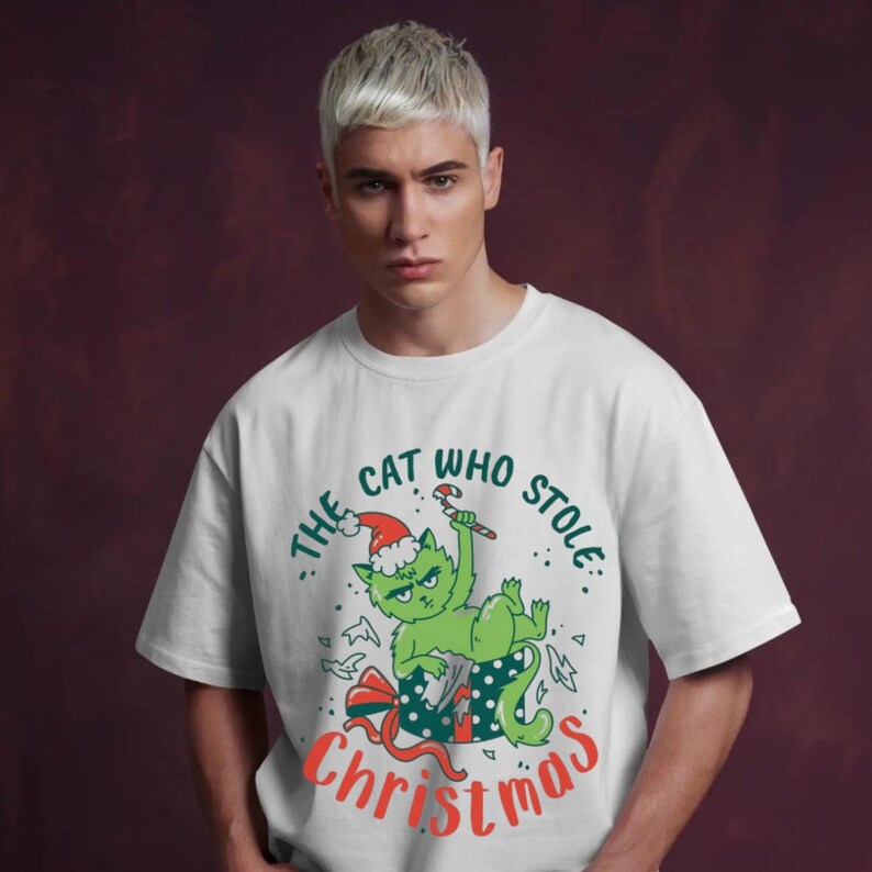 Peut inclure: T-shirt blanc avec un chat vert portant un bonnet de P&egrave;re No&euml;l et tenant un sucre d'orge. Le texte "The Cat Who Stole Christmas" est imprim&eacute; en vert et rouge. Le chat est dans un coffret cadeau avec un n&oelig;ud rouge.