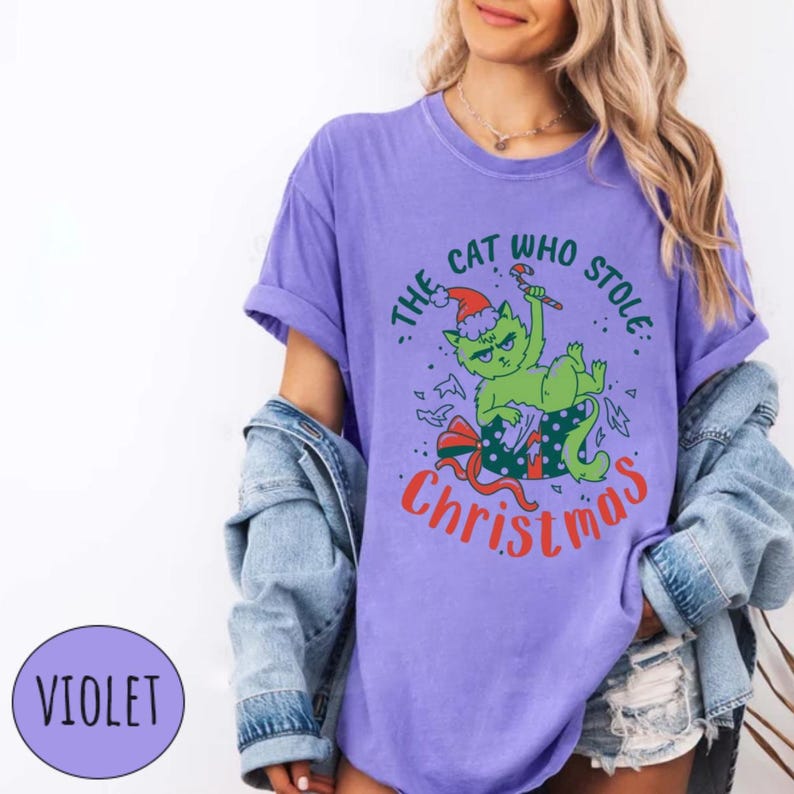 Peut inclure: T-shirt violet avec un chat vert portant un bonnet de P&egrave;re No&euml;l et tenant un sucre d'orge. Le texte dit "The Cat Who Stole Christmas" en vert et rouge. Th&egrave;me de No&euml;l.