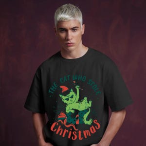 Peut inclure: T-shirt noir avec un chat vert portant un bonnet de P&egrave;re No&euml;l et tenant un sucre d'orge. Le texte "The Cat Who Stole Christmas" est en vert et rouge. Le chat est sur un cadeau emball&eacute;. Design festif.