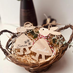 K&ouml;nnte beinhalten: Ein Weidenkorb enth&auml;lt zwei h&ouml;lzerne Hasen-Dekorationen. Jedes Ornament ist mit einem Namen, "Simon" und "Emilia", graviert und mit rot-wei&szlig;em Garn gebunden. Der Korb ist mit Holzsp&auml;nen gef&uuml;llt und mit Gr&uuml;nzeug verziert.