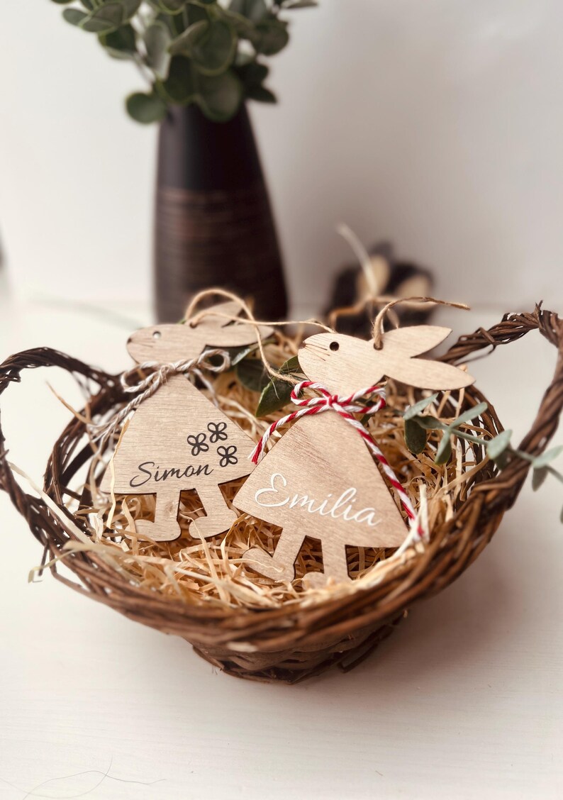 K&ouml;nnte beinhalten: Ein Weidenkorb enth&auml;lt zwei h&ouml;lzerne Hasen-Dekorationen. Jedes Ornament ist mit einem Namen, "Simon" und "Emilia", graviert und mit rot-wei&szlig;em Garn gebunden. Der Korb ist mit Holzsp&auml;nen gef&uuml;llt und mit Gr&uuml;nzeug verziert.