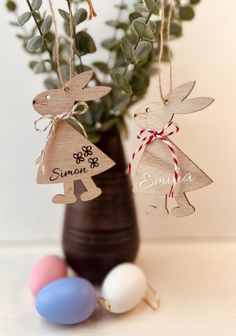 K&ouml;nnte beinhalten: Zwei h&ouml;lzerne Hasenornamente mit Namen und floralen Akzenten h&auml;ngen an einer Schnur. Eines tr&auml;gt den Namen "Simon" und das andere "Emilia". Sie sind mit rot-wei&szlig;en Bindf&auml;den verziert. Pastellfarbene Ostereier sind im Vordergrund.