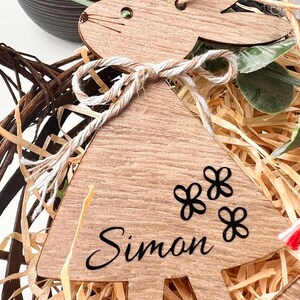 K&ouml;nnte beinhalten: Osterhase-Ornament aus Holz mit dem Namen "Simon" und drei Blumendesigns. Das Ornament ist mit Bindfaden gebunden und sitzt in einem Korb mit Stroh.