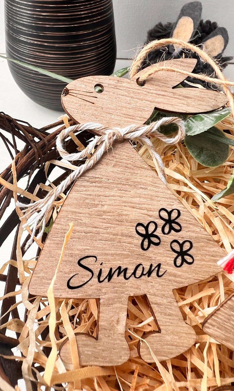 K&ouml;nnte beinhalten: Osterhase-Ornament aus Holz mit dem Namen "Simon" und drei Blumendesigns. Das Ornament ist mit Bindfaden gebunden und sitzt in einem Korb mit Stroh.