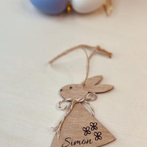 K&ouml;nnte beinhalten: Osterhase-Ornament aus Holz mit dem Namen "Simon" und Blumendetails. Der Hase h&auml;ngt an einer Schnur. Ostereier in Pastellfarben befinden sich im Hintergrund.