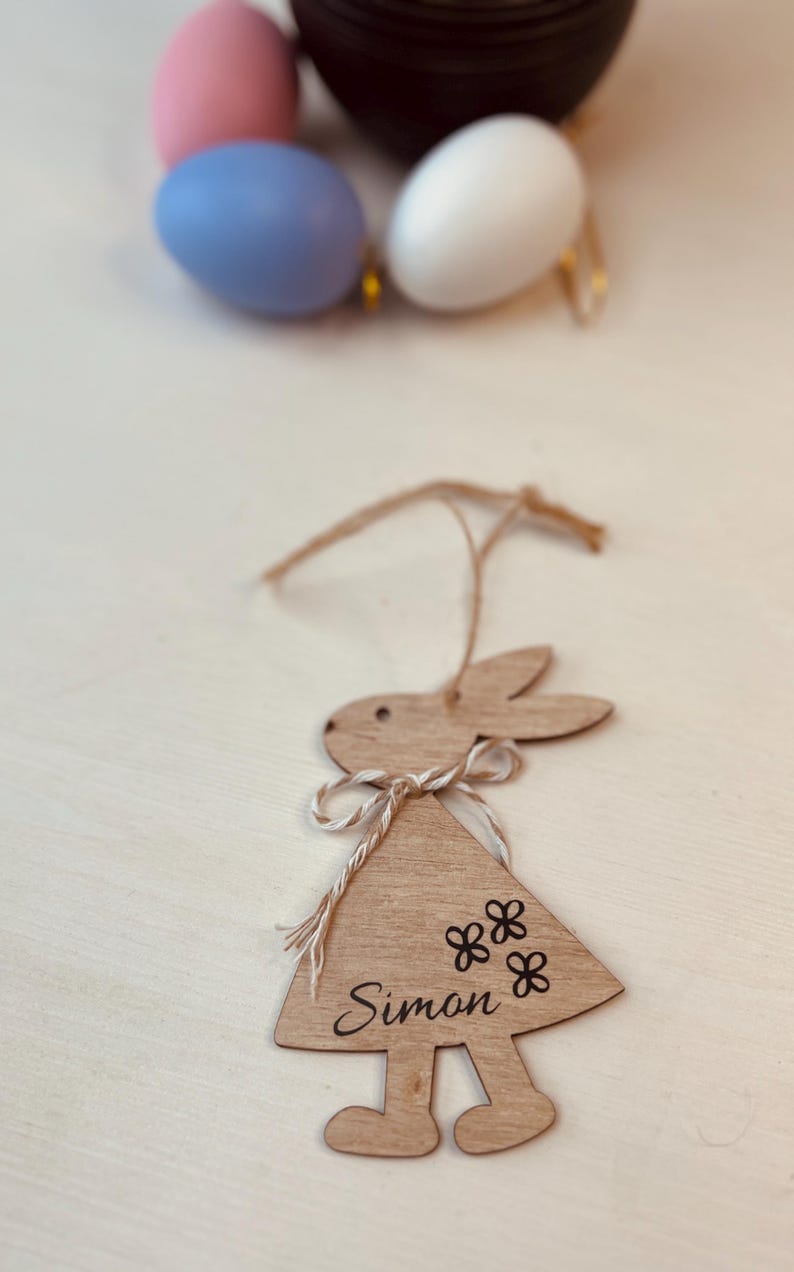 K&ouml;nnte beinhalten: Osterhase-Ornament aus Holz mit dem Namen "Simon" und Blumendetails. Der Hase h&auml;ngt an einer Schnur. Ostereier in Pastellfarben befinden sich im Hintergrund.